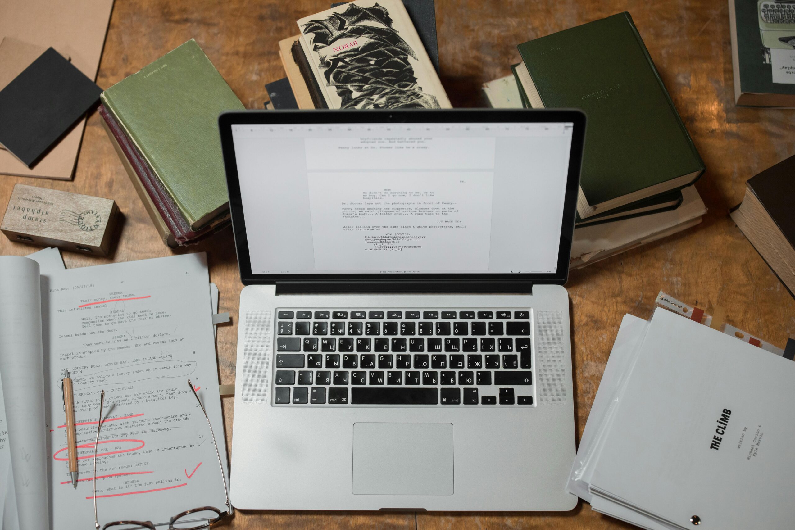 A laptop surrounded by books and scripts on a wooden desk, perfect for literature and technology themes.